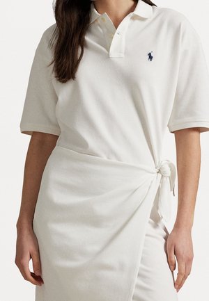 Femme portant une robe polo blanche à manches courtes avec un nœud sur le côté et un petit logo brodé bleu foncé sur la poitrine.