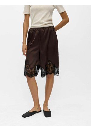 Personne portant un haut beige à manches courtes, un short ample en soie marron foncé avec une bordure en dentelle noire et des chaussures plates noires, debout devant un fond uni.