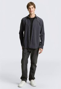 Grijs overhemd met lange mouwen en knopen over een zwart T-shirt, gecombineerd met donkergrijze spijkerbroek en witte sneakers. Glad materiaal, relaxed fit.