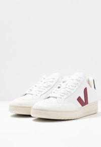 Zapatillas blancas con una suela de goma texturizada, que presentan un logotipo en forma de "V" en color marrón, agujetas a juego y un acento beige en el talón. Diseño minimalista.