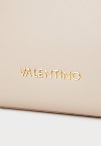 Borsa in pelle beige con logo "VALENTINO" in oro a rilievo. Texture liscia con un design minimalista e senza ulteriori motivi o decorazioni.