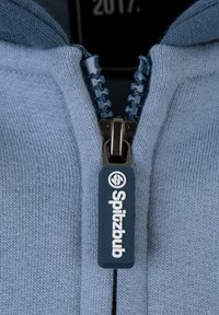 Primo piano di un maglione azzurro chiaro con zip anteriore, dotato di un cursore blu scuro con il logo "Spitzbub" e un tessuto a maglia testurizzato.