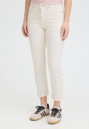 Mujer vistiendo jeans de pierna recta color marfil y zapatillas beige con rayas negras, de pie contra un fondo blanco liso.