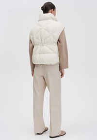 Puffige cremefarbene Weste mit hohem Kragen und elastischem Bund, kombiniert mit hellbeigen Hosen. Glatte Textur mit rautenförmigem Steppmuster.