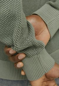 Grauer strukturierter Pullover mit einem erhabenen Muster am Körper und gerippten Bündchen. Der Stoff wirkt weich und leicht dehnbar.