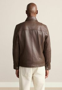 Barna bőrkabát klasszikus gallérral, látható varrással és fémből készült gombokkal. A fazon illeszkedő silhuettet és sima textúrát kínál.