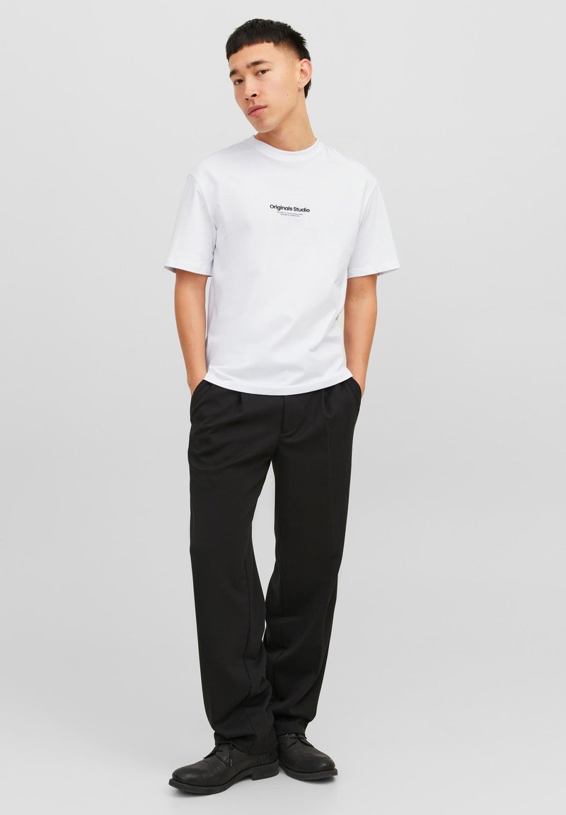 White short-sleeve t-shirt with black text logo, paired with black trousers and black shoes, model standing with hands in pockets.