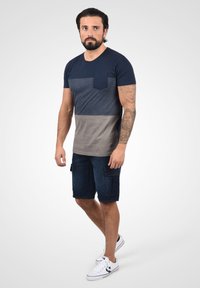 Camiseta a rayas en colores azul marino y gris con un bolsillo en el frente, combinada con pantalones cortos cargo de mezclilla oscura. El modelo lleva zapatillas blancas.