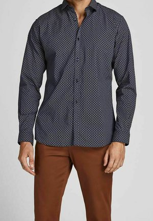Mann trägt ein langärmeliges dunkelblaues Hemd mit kleinem geometrischem Muster und braune Hose, steht mit entspannten Armen.