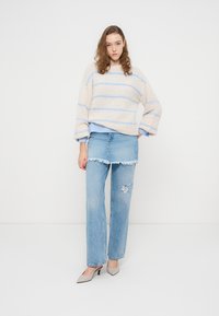 Νεαρή γυναίκα που φοράει πουλόβερ με ρίγες σε μπεζ και ανοιχτό μπλε, στρωμένο τζιν φούστα πάνω από σκισμένα τζιν και μπεζ τακούνια, στέκεται μπροστά σε λευκό φόντο.