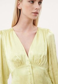 Hellgelbe Satinbluse mit tiefem V-Ausschnitt, gerafften Schultern und Knopfverschluss. Verfügt über ein dezentes Blumenmuster und eine glatte Textur.