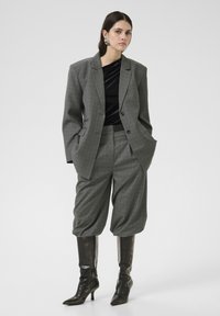 Grauer karierter oversized Blazer mit passenden cropped Hosen. Getragen mit einem schwarzen strukturierten Oberteil und schwarzen kniehohen spitz zulaufenden Stiefeln. Silberne Ohrringe.