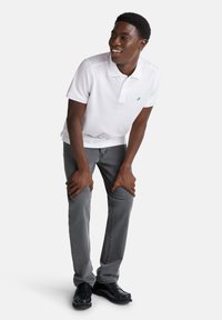 Camisa polo blanca con logo verde, jeans grises y mocasines negros. La persona está de pie y se inclina ligeramente hacia adelante, sonriendo.