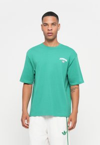 Zöld pamut póló rövid ujjakkal és kerek nyakkal. Bal mellen pink "ADIDAS" logó és háromlevelű lóhere található.