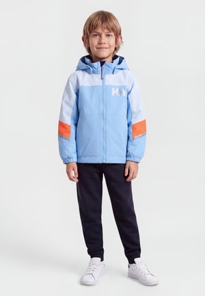 Barn iført en lyseblå og hvid jakke med orange striber på ærmerne, mørke bukser og hvide sneakers, stående vendt mod fronten.