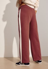 Mørkerøde joggingbukser med hvide sidelommer, afslappet pasform, elastisk taljebånd og blød tekstur. Brugt med mørke sneakers.