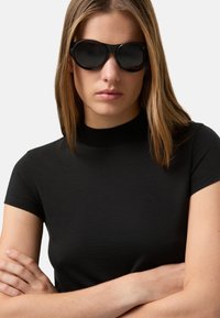 Černý krátký rukáv z hladkého materiálu, doplněný o velké černé sluneční brýle s kulatým designem a neprůhlednými čočkami.