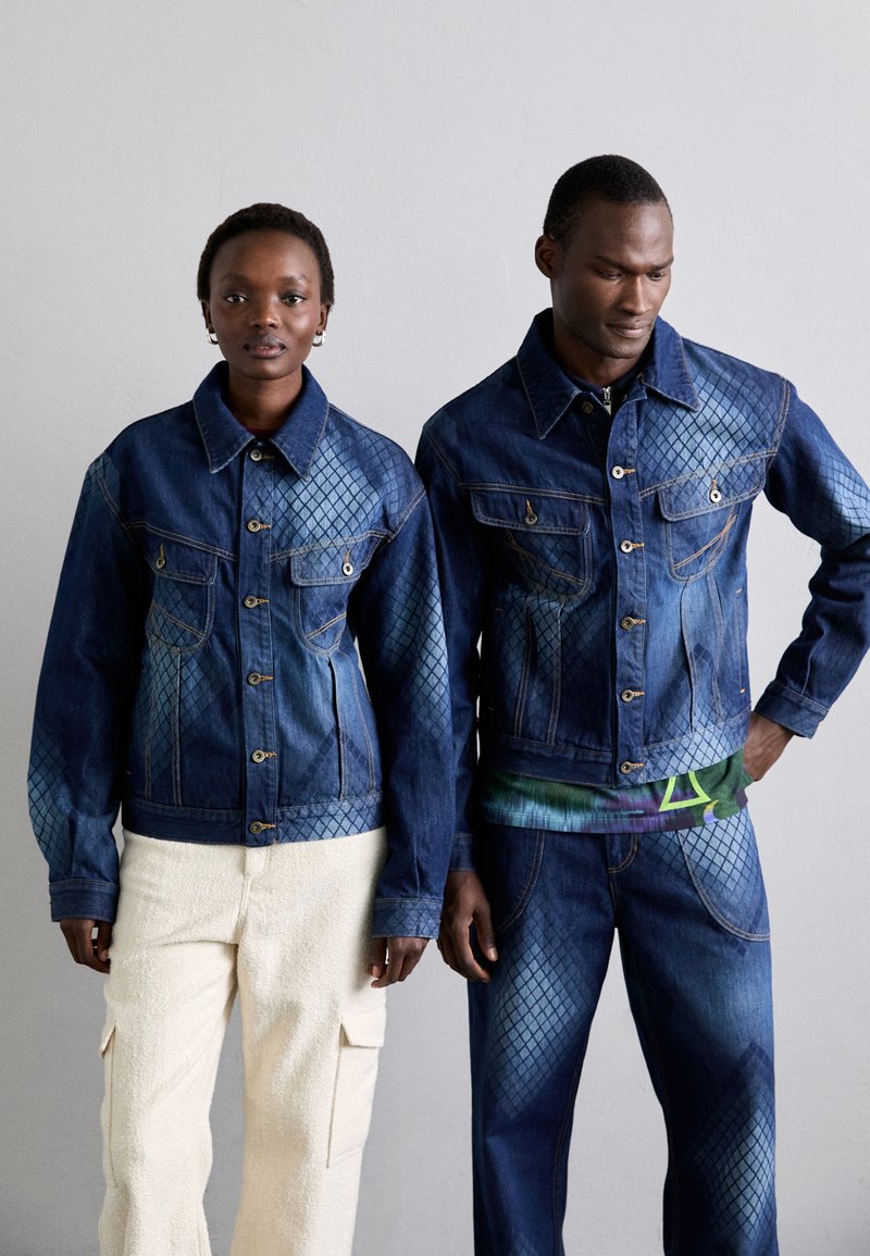 Two models wear matching blue denim jackets with diamond-patterned accents, featuring button closures and chest pockets. The female model pairs hers with cream pants.