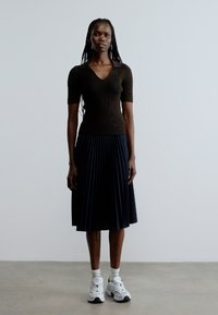 Top marrón acanalado de manga corta, falda midi plisada azul marino, zapatillas blancas, llevado por una figura de pie sobre un fondo claro.