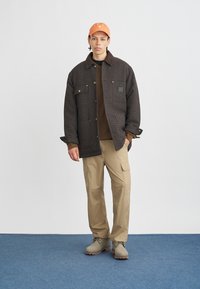 Giacca a quadri marrone con grandi bottoni, maglia marrone, pantaloni cargo color sabbia e stivali beige. Il berretto arancione aggiunge un dettaglio a contrasto.