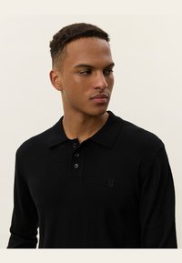 Zwarte longsleeve polo met een kraag, drie-knopen sluiting en geborduurd logo op de borst. Glad textuur en getailleerd design.
