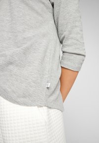 Graues langärmliges Oberteil mit weicher Textur, abgerundetem Saum und seitlichem Etikett, kombiniert mit weißen gesteppte Hosen, die eine sichtbare Tasche haben.