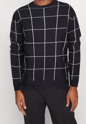 Homme portant un pull noir à motif quadrillé blanc et un pantalon noir, debout devant un fond clair uni.