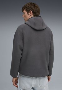 Grauer Fleece-Hoodie mit passender Kapuze, überschnittenen Schultern und weicher Textur. Präsentiert ein minimalistisches Design und eine lockere Passform.