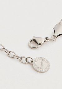 Αλυσίδα από ασημένιο μέταλλο με κούμπωμα αχινός, λεία υφή και στρογγυλή ετικέτα με ενσωματωμένες λεπτομέρειες. Απλός, λειτουργικός σχεδιασμός.