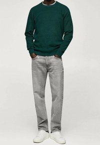 Personne portant un pull vert foncé, un jean gris clair et des baskets blanches, debout les mains dans les poches devant un fond uni.