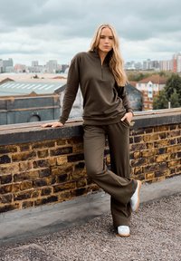 Blonde vrouw in olijfgroene ritsvest en broek leunt op een stenen dakrand met op de achtergrond stadsgebouwen en een bewolkte lucht.