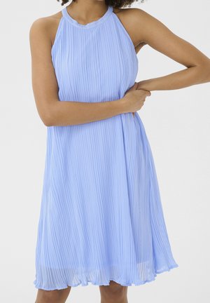Žena v světlomodrých plisovaných šatech bez rukávů, držící se za bok s překříženýma rukama na jednobarevném pozadí.