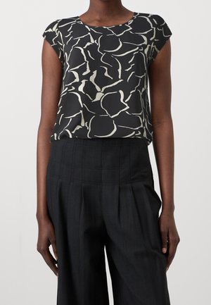 Kvinde iført en sort bluse med korte ærmer og hvidt abstrakt print, stoppet ned i sorte, højtaljede, plisserede bukser.