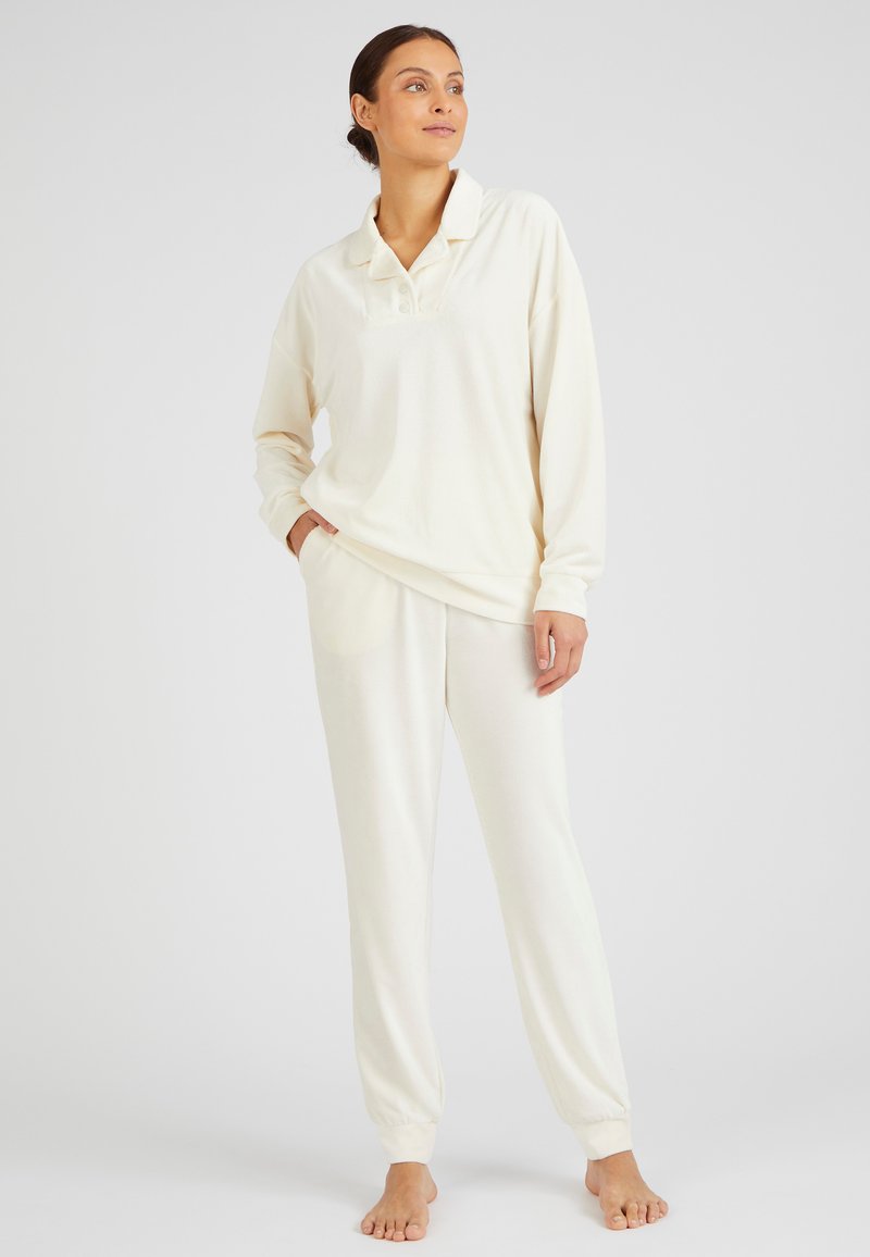 El conjunto de loungewear de color crema presenta un pulóver con cuello y botones, junto con joggers con cintura elástica, fabricados con un tejido suave y texturizado.