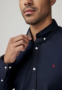 Camicia blu navy con chiusura a bottoni e finitura testurizzata, colletto a contrasto a quadri. Presenta un piccolo logo rosso sul petto e bottoni bianchi.