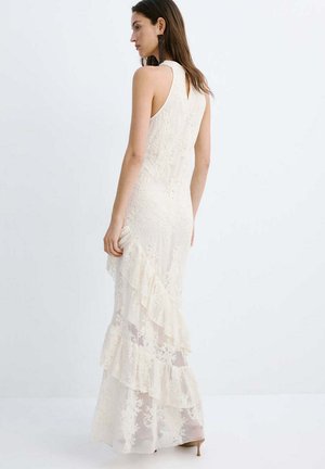 Mujer con un vestido de encaje crema sin mangas y volantes superpuestos, de perfil sobre un fondo blanco liso.