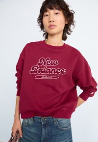 Vynikanti bordo spalvos megztinis su baltu "New Balance Athletics" logotipu, apvaliu kaklu ir ruoželinėmis rankovėmis, derinamas su mėlynomis džinsinėmis kelnėmis.