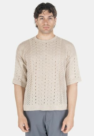 Giovane uomo che indossa un maglione beige a maniche corte con buchi decorativi e pantaloni grigi, in piedi fronteggiando su sfondo bianco.