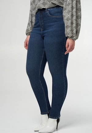 Skinny jeans van donkerblauwe denim, met een hoge taille, subtiele stiksels en een gladde textuur, gecombineerd met witte enkelboots.