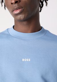 Ανοιχτό μπλε φούτερ με ραμμένο στρογγυλό λαιμό και λευκό λογότυπο "BOSS" στο μπροστινό μέρος. Λείο υλικό, εφαρμοστό σχέδιο.