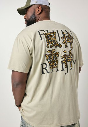 Uomo che indossa una T-shirt beige con testo nero e caratteri asiatici stampati con motivo leopardato, cappellino verde e bianco, in piedi davanti a uno sfondo uniforme.