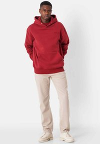 Sudadera roja con capucha, con un bolsillo frontal y puños acanalados, combinada con pantalones de color claro y zapatos blanco roto sobre un fondo neutro.