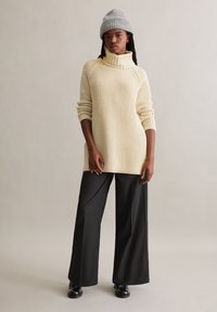 Cremiger Strickpullover mit hohem, umgeschlagenem Kragen, kombiniert mit weit geschnittenen schwarzen Hosen und schwarzen Stiefeletten, vor einem neutralen Hintergrund.