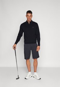 Sweatshirt noir zippé, shorts sombres et baskets blanches. Le mannequin tient un club de golf, avec un fond gris uni qui met en valeur les détails de la tenue.