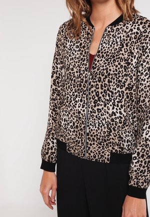 Vrouw draagt een ritsvest met luipaardprint, zwarte manchetten en zoom, gecombineerd met zwarte broek tegen een effen achtergrond.