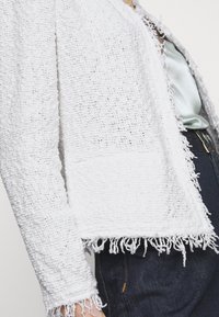 Biała krótka kurtka wykonana z teksturowanej tkaniny tkanej, z ozdobnym frędzlem na krawędziach, o dopasowanym kroju i otwartym przodzie.