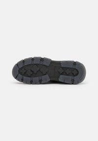 Semelle de botte en caoutchouc noir avec des sculptures profondes et des motifs en forme de croix pour l’adhérence sur un fond gris clair.