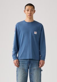 Langärmliges blaues Baumwollhemd mit einer Brusttasche, bedrucktem Text auf dem Ärmel und einem lässigen Schnitt. Getragen mit leichter Denim.