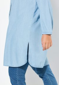 Tunic in denim azzurro con orlo curvo, maniche lunghe e spacchi laterali, indossata sopra leggings a pois blu scuro. Texture morbida.