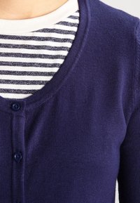 Cardigan azul marino hecho de material de punto, con cuello en V, cierre de botones, y llevado sobre una camisa a rayas blancas y azules.